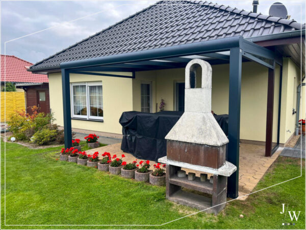 Moderne anthrazitfarbene Terrassenüberdachung aus Aluminium an einem Einfamilienhaus mit gepflegtem Garten, roten Blumenbeeten und einem freistehenden Außenkamin