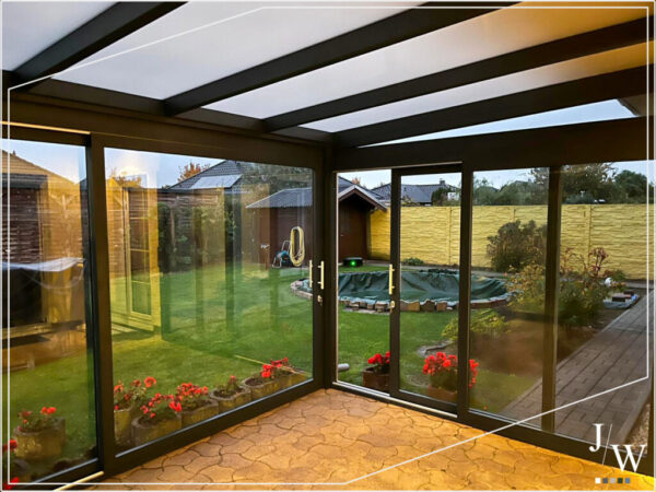 Verglaste Terrassenüberdachung mit großen Schiebetüren und Blick in einen gepflegten Garten mit Rasen, Blumenbeeten und Gartenhaus