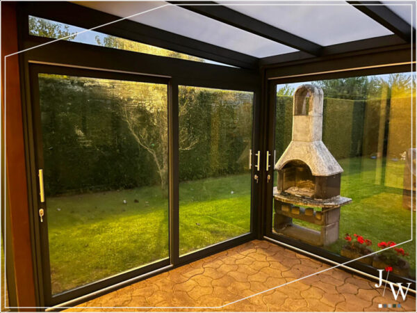Geschlossene Terrassenüberdachung mit bodentiefen Glaswänden und Sicht auf einen Außenkamin sowie den umliegenden Garten