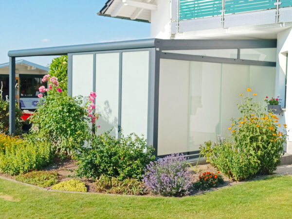Seitlich geschlossene Terrassenüberdachung mit opalem Polycarbonat und grauer Aluminiumkonstruktion, umgeben von blühendem Garten